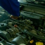 Mechanic in blue uniform working on a car engine in a garage environment.